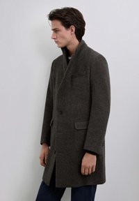 Abrigo marrón en espiga con cuello de muesca, cierre de un botón y bolsillos laterales. Corte entallado con hombros estructurados, textura de mezcla de lana.