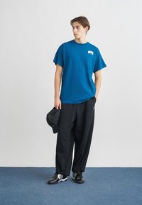 Μπλε βαμβακερό t-shirt με κοντά μανίκια και ένα μικρό γραφικό, σε συνδυασμό με μαύρο παντελόνι φαρδύ και μαύρα αθλητικά παπούτσια. Μαύρο καπέλο κρατημένο στο χέρι.