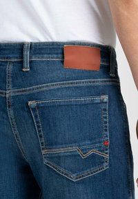 Dunkelblaue Jeans aus Denim mit einem braunen Lederpatch auf der Rückseite, gelben Nähten, seitlichen und hinteren Taschen sowie einem roten Akzent auf der Hintertasche.