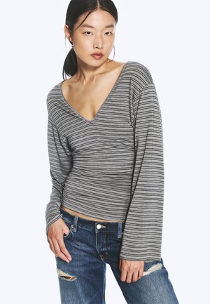 Jeune femme aux cheveux noirs attachés porte un t-shirt rayé gris à manches longues et col en V, ainsi qu’un jean bleu déchiré, debout avec une main dans la poche.