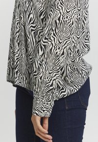 Černobílá pruhovaná košile se vzorem zebry, dlouhými rukávy a volným střihem, kombinovaná s tmavě modrými džíny.