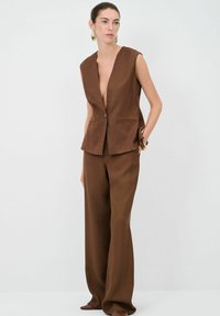 Brauner ärmelloser Blazer mit tiefem V-Ausschnitt, einem Knopfverschluss und zwei Fronttaschen, kombiniert mit passenden weiten Hosen. Glatte Stoffstruktur.
