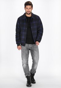 Karierte Jacke in Marine- und Blautönen, schwarzes Rundhalshemd, graue Hose und schwarze Schnürstiefel. Mit Knöpfen und Kragen.