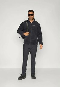 Črna flis jakna z zadrgo, v kombinaciji s črnimi hlačami in planinskimi čevlji. Model nosi črn nahrbtnik in ima temne sončne očala.