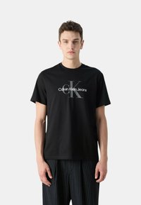 Camiseta de algodón negra con un logo en blanco y gris de "Calvin Klein Jeans". Mangas cortas, escote redondo, combinada con pantalones a rayas.