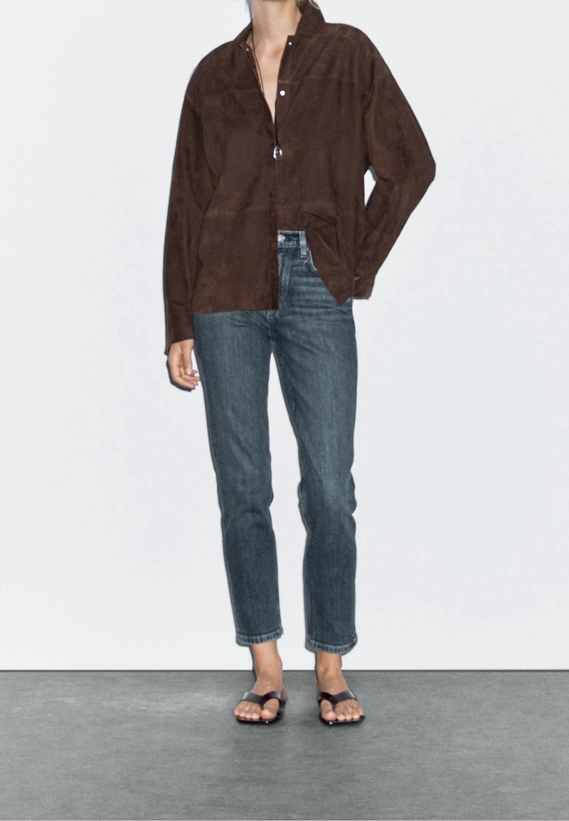 Personne portant une chemise marron foncé à manches longues, un jean bleu droit et des sandales noires à bout ouvert, debout sur un sol gris contre un mur blanc.