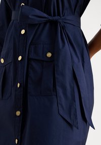 Temno modra obleka z zlatimi gumbi, dvema sprednjima žepoma s poklopcem in pasom iz tkanine, ki je zavezan v pentljo na pasu.