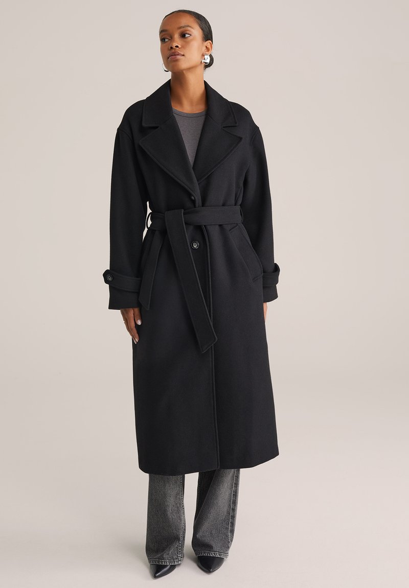Donna che indossa un lungo cappotto nero con cintura sopra un top grigio e jeans neri sbiaditi, in piedi con le mani rilassate ai lati su uno sfondo neutro.