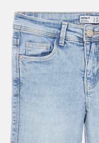 Ανοιχτό μπλε τζιν παντελόνι με ξεθωριασμένο πλύσιμο, που διαθέτει μικρή μπροστινή τσέπη, μεταλλικό κουμπί κλεισίματος και ραμμένες λεπτομέρειες στις ραφές.