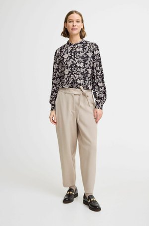 Bluse mit floralem Druck in Schwarz und Beige, langärmlig und mit Knopfdetails, kombiniert mit beigen, hoch taillierten Hosen und schwarzen Loafers.