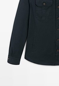 Mørkeblå skjorte med lange ærmer, knaplukning ved manchet, knapper foran, brystlomme med klap og sidelomme på hvid baggrund.