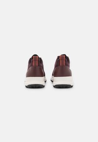 Bakvy av sneakers med en vinröd tygöverdela, brun syntetisk häl, texturerad burgunderröd dragflik och en vit mellansula med svart yttersula.