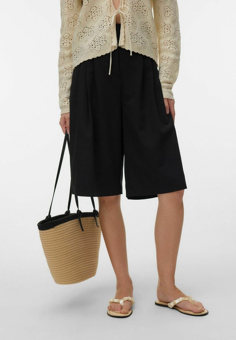 Personne portant un cardigan beige en crochet, un short noir jusqu'aux genoux, tenant un sac seau en paille et portant des sandales ornées de perles.