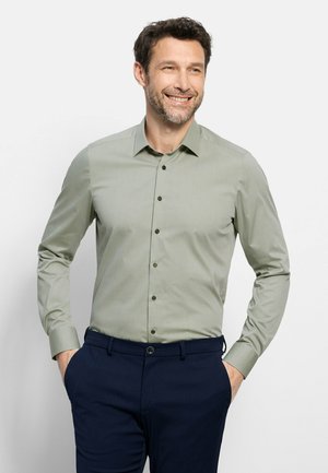 Glimlachende man met handen in de zakken, gekleed in een lichtgroen overhemd en donkerblauwe broek tegen een effen lichte achtergrond.