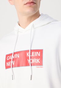 Hanorac alb cu un imprimeu grafic cu dreptunghiuri roșii. Textul include "CALVIN NEW KLEIN YORK." Șnururile au capete argintii.