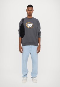 Grijze sweatshirt met een wit en oranje grafisch logo, lichtblauwe oversized jeans en witte sneakers. Zwarte tas over de schouder.