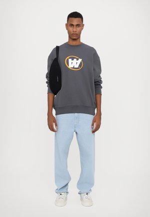 Gray sweatshirt with a white and orange graphic logo, light blue oversized jeans, and white sneakers. Black bag slung over the shoulder.
