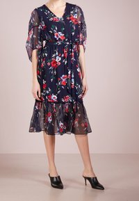 Mujer que lleva un vestido midi floral azul marino con mangas abullonadas transparentes y dobladillo, con cintura ajustada, combinado con mules negros de tacón y punta.