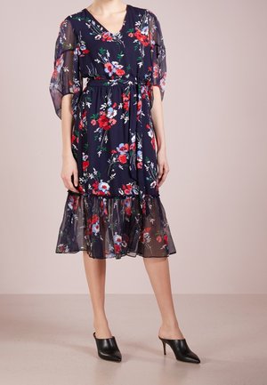 Femme portant une robe midi florale marine avec des manches bouffantes transparentes et un ourlet, taille cintrée, associée à des mules à talons aiguilles noires.