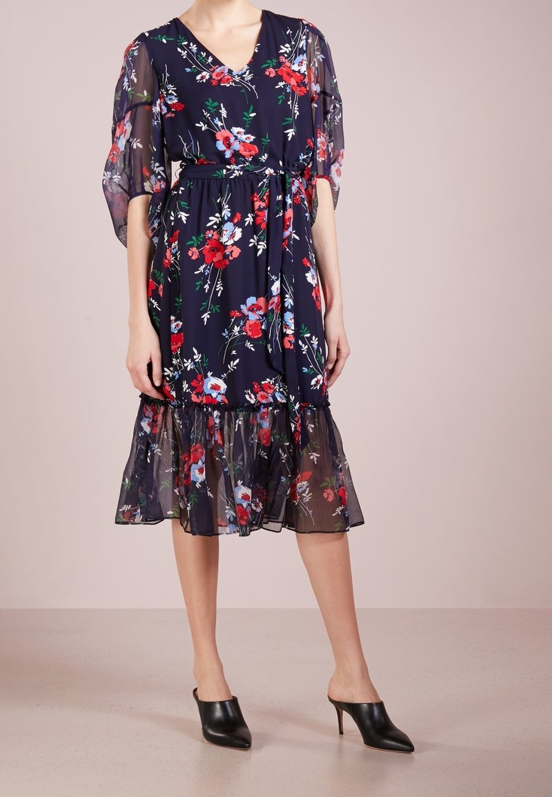 Mujer que lleva un vestido midi floral azul marino con mangas abullonadas transparentes y dobladillo, con cintura ajustada, combinado con mules negros de tacón y punta.