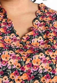 Primo piano di un colletto di una blusa floreale multicolore con bordi ondulati, caratterizzata da fiori arancioni, rosa e rossi su uno sfondo scuro.