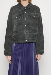 Svart denimjacka med en kortad passform, knäppning framtill, två framfickor och kontrasterande sömmar, ihop med en veckad lila kjol.