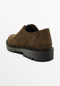 Chaussure en daim brun avec une épaisse semelle en caoutchouc noir, présentant un bout arrondi et un design minimal sans lacets ni éléments apparents.