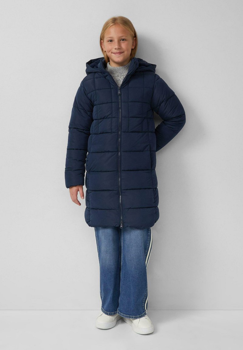 Donkerblauwe gewatteerde jas met capuchon, voorzien van een quiltpatroon, ritssluiting aan de voorkant en lange mouwen, gedragen over een jeans in het blauw en witte sneakers.
