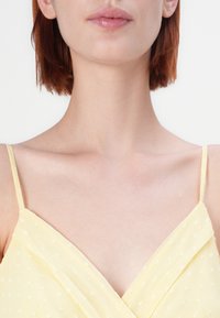 Gul, ærmeløs kjole med indpakningsdesign. Stoffet har en subtil tekstur og små firkantede mønstre overalt.