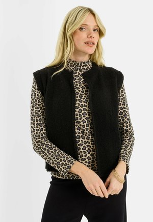 Mujer rubia con chaleco de forro polar negro sin mangas sobre una blusa de manga larga con estampado de leopardo, y pulseras doradas en la muñeca, sobre fondo blanco.