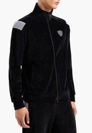 Giacca nera in velluto a coste con zip, caratterizzata da accenti a strisce sulle spalle e un logo patch discreto. Include tasche laterali e un colletto alto.