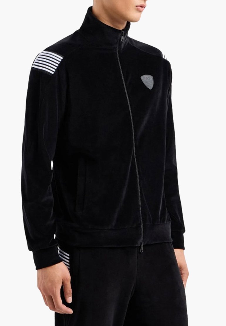 Giacca nera in velluto a coste con zip, caratterizzata da accenti a strisce sulle spalle e un logo patch discreto. Include tasche laterali e un colletto alto.