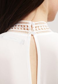 Biała bluzka z wysokim dekoltem, ozdobiona koronkowym wykończeniem i dwoma guzikami pokrytymi tkaniną z tyłu. Gładka faktura i elegancki design.