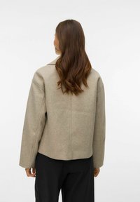 Bēšs vilnas maisījuma žakete ar brīvu piegriezumu, lielu apkakli un nolaistām pleciem; attēlots no aizmugures ar gludu tekstūru un minimālu dizainu.