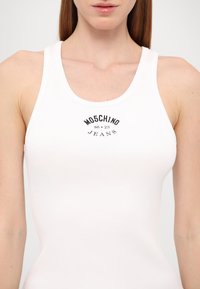 Débardeur blanc côtelé avec un col rond et des bretelles épaisses, présentant une impression graphique noire indiquant "MOSCHINO JEANS" au-dessus du centre.