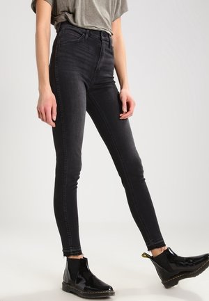 Personne portant un jean skinny noir taille haute avec des ourlets effilochés, un t-shirt gris rentré, et des bottines noires brillantes sur un fond clair.