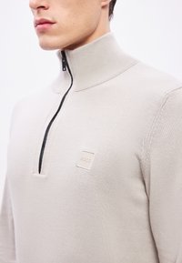 Beeži poollukuga kampsun soonikkoelise kraega, millel on väike kreemjas ruudukujuline plaastrik sildiga „BOSS“ ja kena tekstuuriga kudumimuster kogu pinnal.