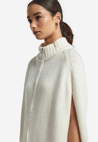 Vit stickad cape med hög krage, med dragkedja fram och sidofickor. Mjuk textur med ribbade detaljer på kragen och kanterna.