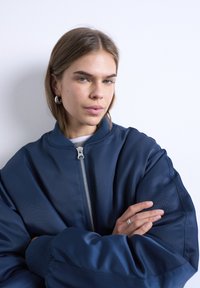 Νεαρή γυναίκα με ανοιχτό καστανά μαλλιά, ντυμένη με μπλε σκούρο μπουφάν με φερμουάρ, ασημένια στεφάνια στα αυτιά και ασημένιο δαχτυλίδι, στέκεται με σταυρωμένα τα χέρια.