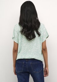 Blusa verde menta dal taglio ampio con un motivo floreale bianco testurizzato. Maniche corte e orlo curvo. Abbinata a jeans blu scuro.
