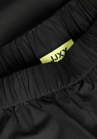 Tissu noir avec une taille élastique froncée et une étiquette de vêtement jaune partiellement visible avec le texte noir "JJXX".