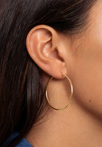 Acercamiento a la oreja de una persona con cabello oscuro que lleva un gran pendiente de aro delgado en oro.