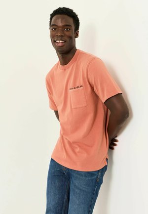 Lächelnder Mann in einem korallenfarbenen Kurzarmhemd mit Tasche und blauer Jeans steht mit den Händen hinter dem Rücken vor einer weißen Wand.