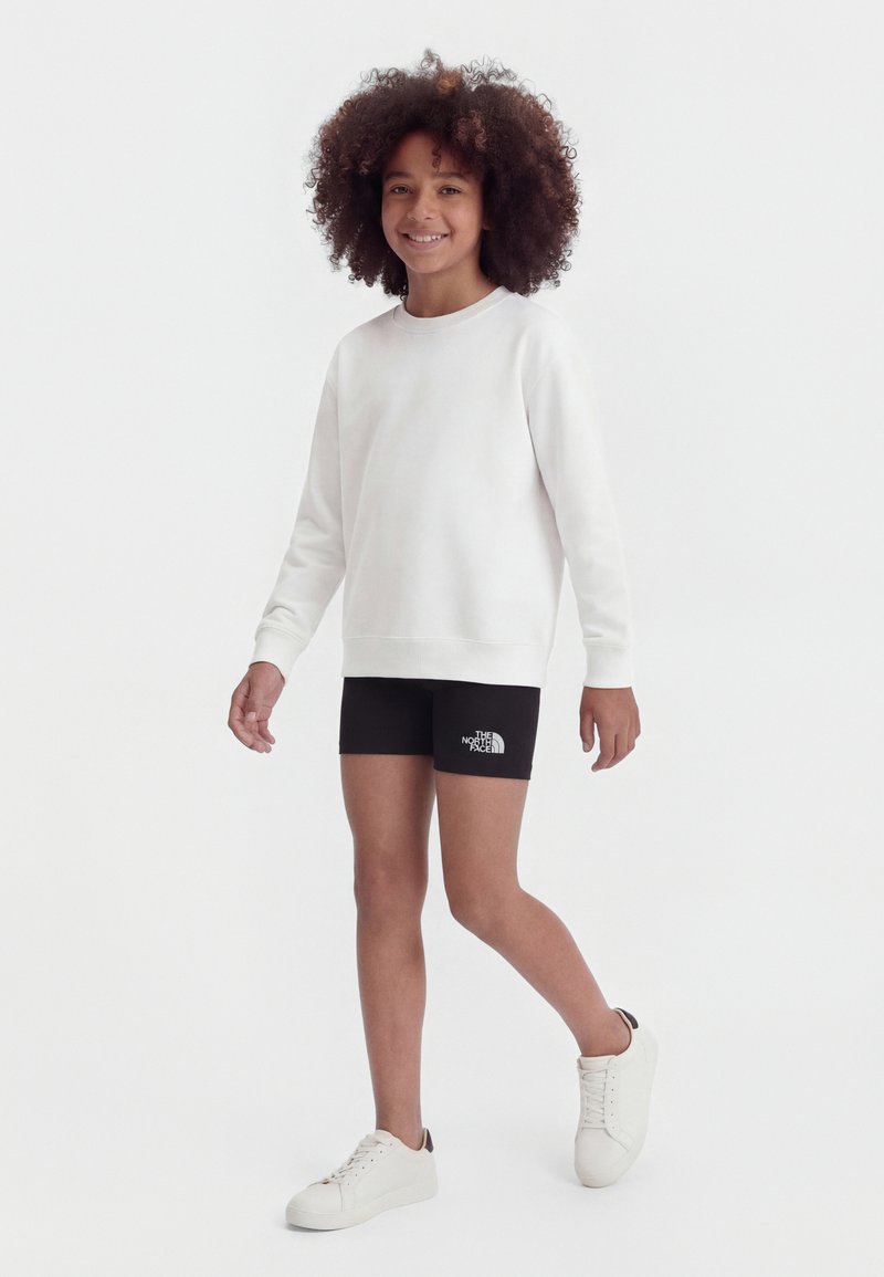 Παιδί με σγουρά μαλλιά, ντυμένο με λευκό φούτερ, μαύρα σορτς The North Face και λευκά αθλητικά παπούτσια, περπατάει και χαμογελάει σε λευκό φόντο.