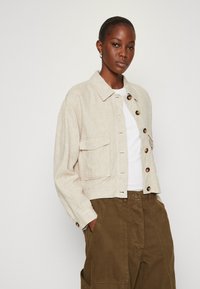 Beige linnen cropped jasje met bruine knopen, twee voorzakken en een kraag, gecombineerd met olijfgroene broek en een witte top.