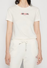 Kvinna i vit kortärmad Armani Exchange T-shirt och matchande dragsko-byxor med röda och svarta logodetaljer.