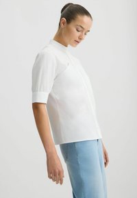 Chemise blanche à manches courtes avec un col haut, dotée d'une fermeture à boutons sur le devant et d'un design ajusté. Portée avec un jean en denim bleu clair.