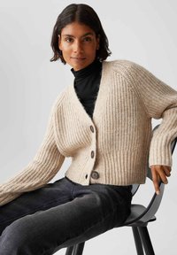Donna con capelli neri corti che indossa un cardigan di maglia beige su un dolcevita nero, jeans scuri, seduta su una sedia nera contro uno sfondo semplice.