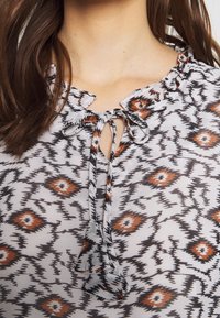 Blouse met een grijze achtergrond en een geometrisch patroon in zwart en bruin. Vastgebonden halslijn met een rucherand, lichte stof.