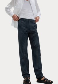 Pantalon bleu foncé à rayures, en tissu, avec une coupe droite, des poches avant et une fermeture à bouton, associé à des sandales noires.